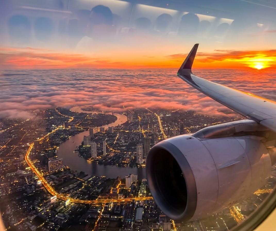 Airplane window view showing the aircraft wing and engine flying over the sprawling city grid of Bangkok during sunset Delhi to Bangkok Flight View